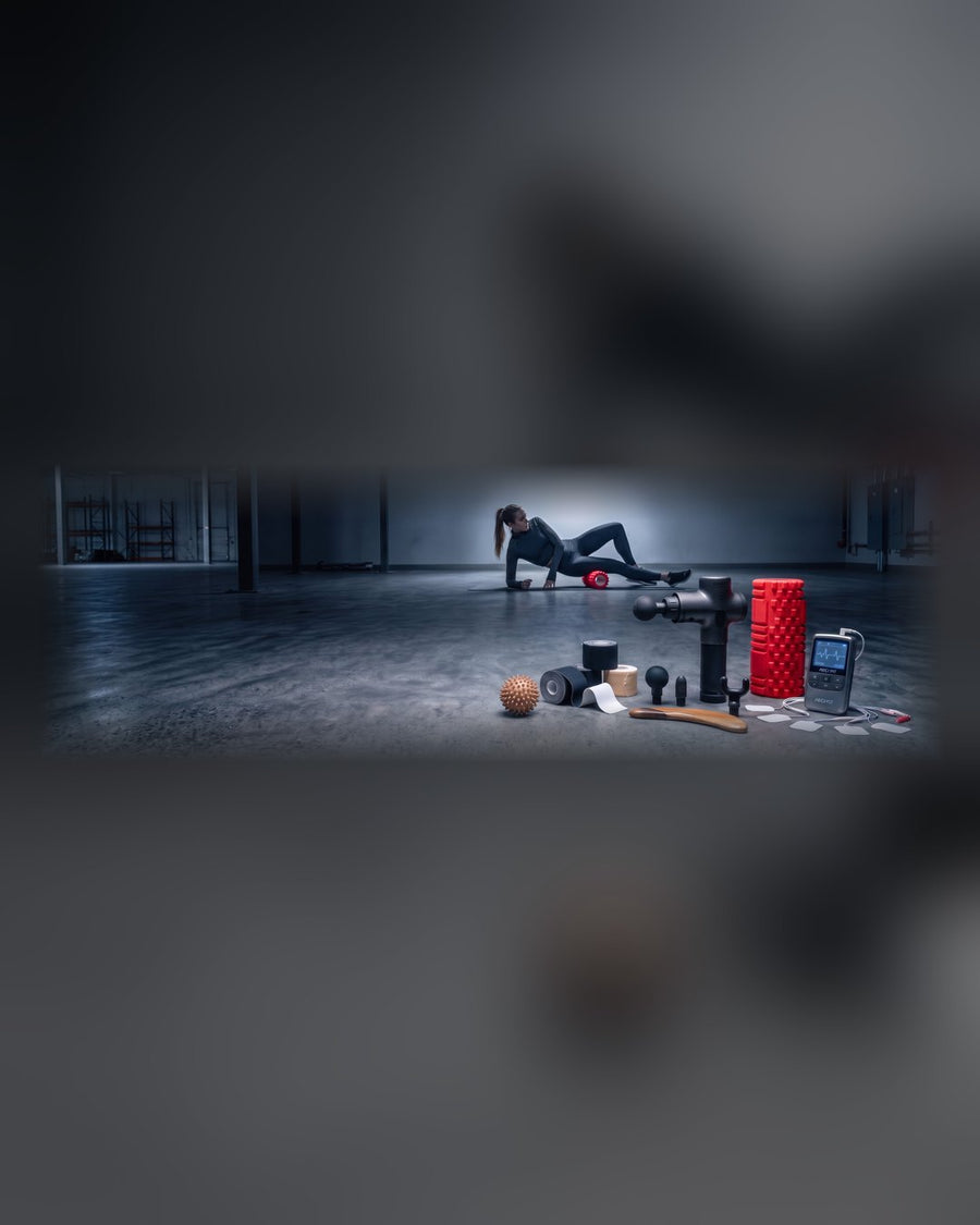 A woman foam rolling on a mat in a cold empty gym, with a massage gun, spiky ball, kinesiology tape, foam roller and a TENS unit set out in front.